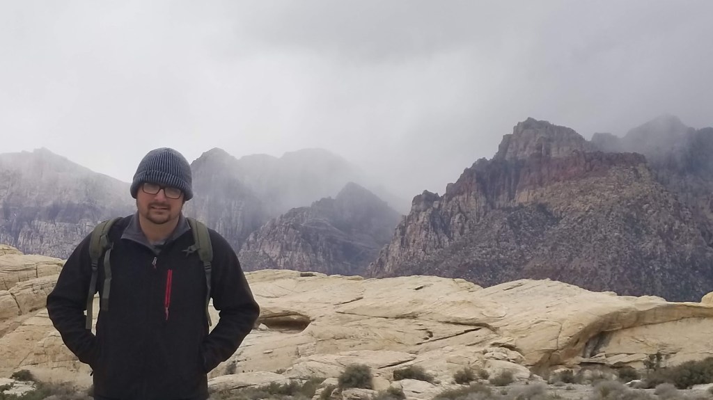 Nate Ballew standing with the Red Rock Canyon behind him. The clouds are pouring over the mountain like a water fall.